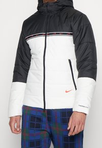 Vadderad jacka med svart övre del och vit undre del. Röd- och svartrandig detalj över bröstet. Har huva och fickor med dragkedja.