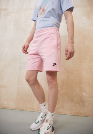 Rosa Baumwollshorts mit elastischem Bund, Seitentaschen und einem schwarzen Nike-Logo. Kombiniert mit einem blauen T-Shirt und weißen Sneakers.