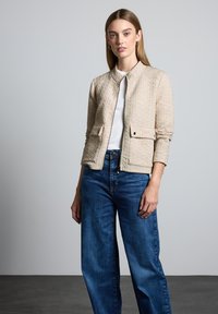 Veste matelassée beige de style bomber, avec une fermeture éclair sur le devant, deux poches avant et un col rond, associée à un jean bleu à jambes larges.