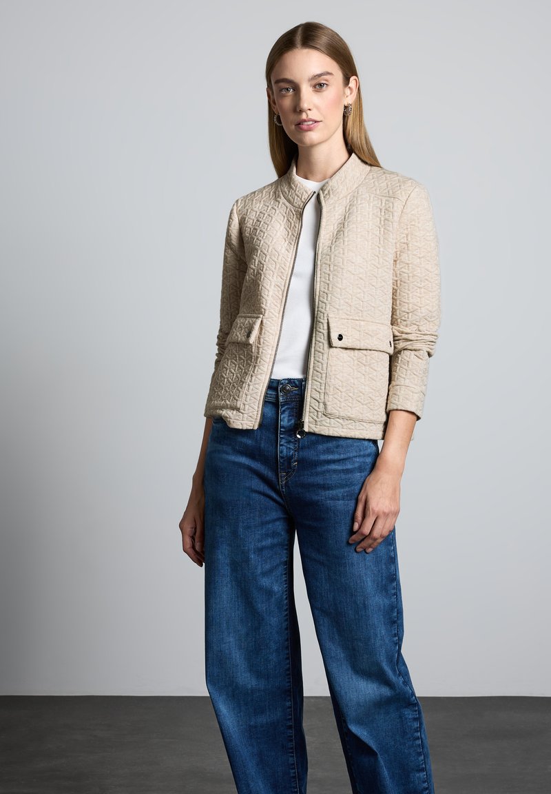 Veste matelassée beige de style bomber, avec une fermeture éclair sur le devant, deux poches avant et un col rond, associée à un jean bleu à jambes larges.