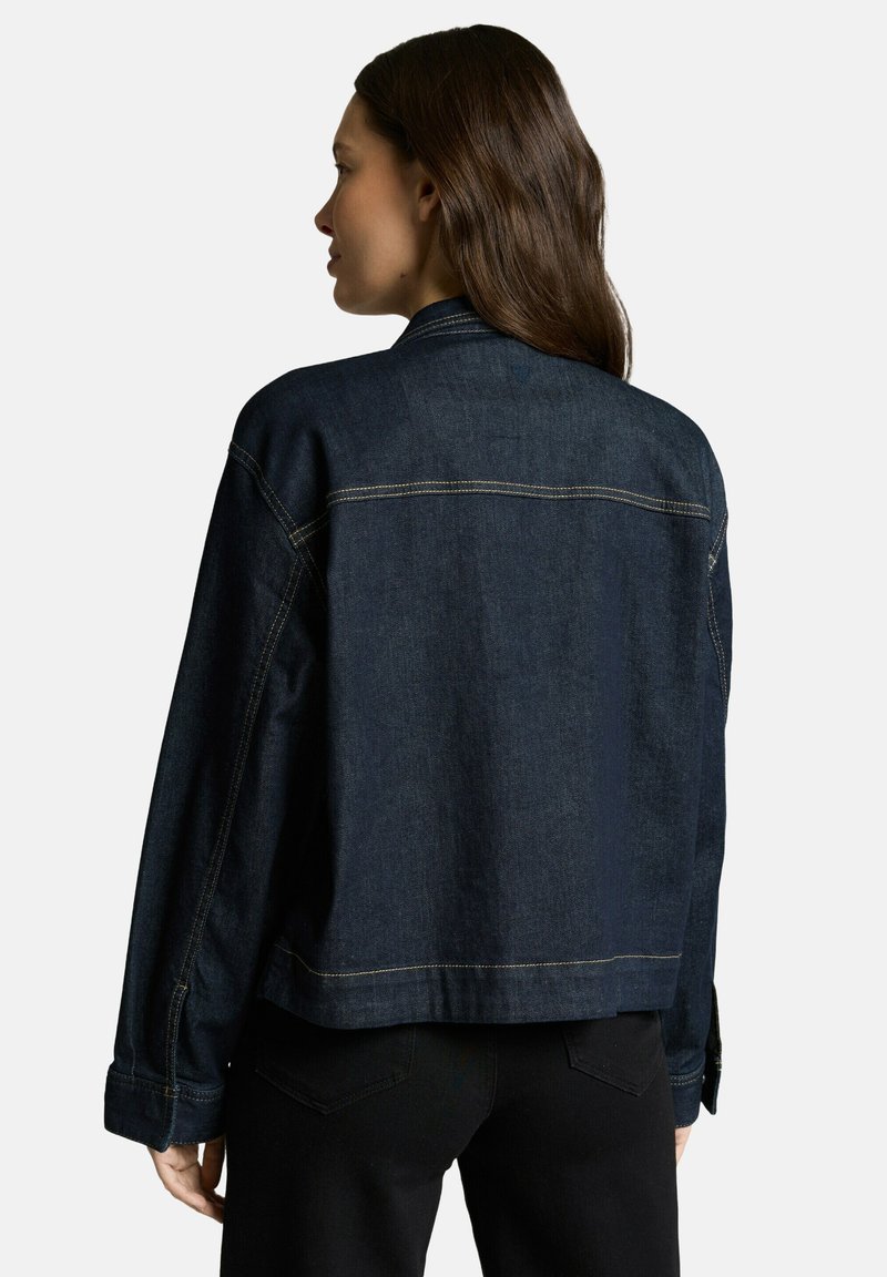 Vrouw met lang donkerbruin haar, gekleed in een donkerblauwe spijkerjack en zwarte broek, met de rug naar de camera tegen een effen lichte achtergrond.