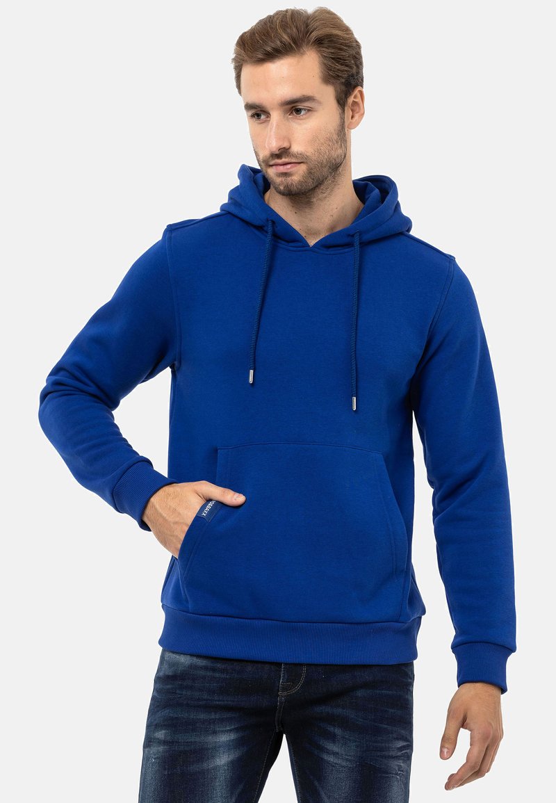 Blauer Hoodie aus weichem Material, ausgestattet mit einer Kängurutasche, verstellbaren Kordeln und gerippten Manschetten. Getragen mit dunklen Jeans.