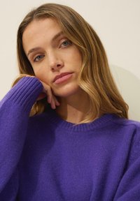 Lila gestrickter Pullover mit rundem Halsausschnitt, gerippten Bündchen und weicher Textur, getragen von einer Person mit hellen, welligen Haaren.
