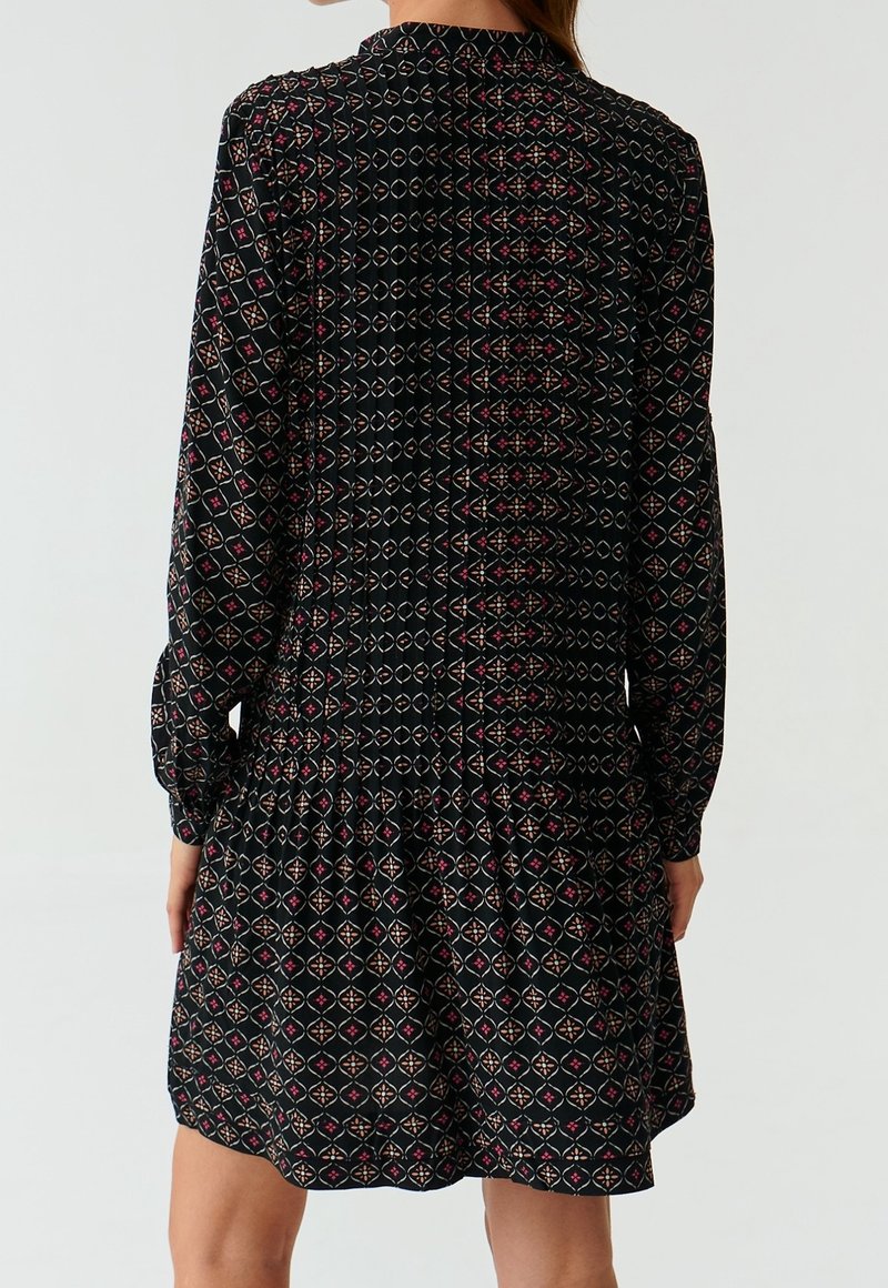 Femme portant une robe noire à manches longues avec un motif géométrique de formes blanches, rouges et orange, vue de dos.