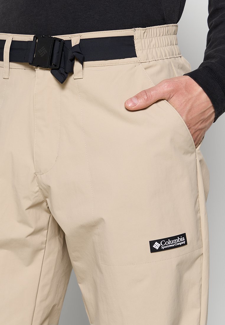 Pantalons légers beiges avec une taille élastique noire, une ceinture ajustable, des poches latérales et un logo Columbia sur la cuisse droite.