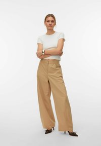 Femme debout, bras croisés, portant un haut blanc à manches courtes, un pantalon beige large taille haute, et des talons pointus marron avec des ornements argentés.