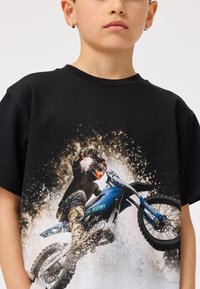 Melna kokvilnas t-kreklu ar motocrossa braucēja grafiku uz zila velosipēda, ar izsplāšanās efektiem un tekstu "Brauc tagad!" zaļā krāsā.