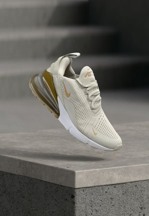 Beżowy but sportowy Nike Air Max z pomarańczowymi logo i widoczną podeszwą z poduszką powietrzną, unoszący się nad betonową krawędzią wewnątrz pomieszczenia.