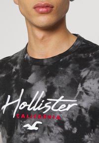 Svart tie-dye t-shirt med rund halsringning. Framsidan har vit broderad text "Hollister" med "CALIFORNIA" i röd färg och en logotypakcent.