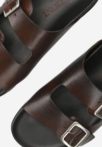 Sandales en cuir marron avec deux brides réglables en métal, design ouvert sur le devant, et une semelle intérieure noire lisse avec un logo.