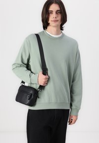 Sort læder crossbody taske med to lynlåslommer, båret over en olivengrøn sweatshirt. modellen poserer afslappet med sorte bukser.