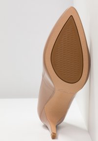 Escarpin nude à talon aigu, avec un bout pointu, une tige en cuir lisse, un talon aiguille fin, et une semelle en caoutchouc texturée arborant un logo de marque.