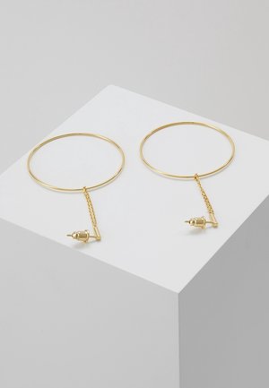 Paire de boucles d’oreilles créoles fines en or avec petite barre verticale et détail de chaîne, présentées sur un cube blanc sur fond gris.