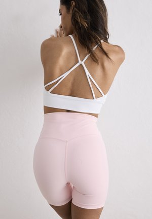 Žena s dlouhými vlasy, oblečená v bílém sportovním topu na tenkých ramínkách a světle růžových vysokým pasem kraťasech, stojící čelem k bílé stěně a dotýkající se jí.