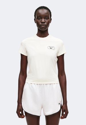 Jeune femme portant un t-shirt blanc ajusté à manches courtes et un short blanc assorti avec un petit logo sur chaque pièce.