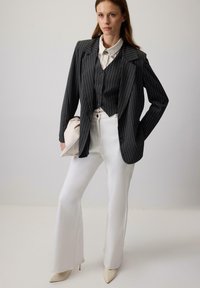 Vrouw in een zwart gestreepte blazer en vest over een witte blouse, witte flared broek, beige hakken, met een beige clutch in de hand, staat zelfverzekerd.