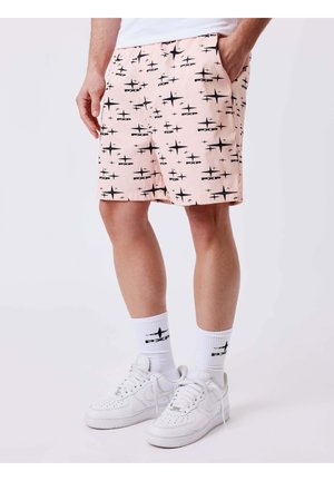 Uomo che indossa pantaloncini rosa chiaro con motivi a stelle nere, calzini bianchi a metà polpaccio con logo nero e scarpe da ginnastica bianche, in piedi con una mano in tasca.