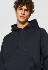 Felpa blu navy con tasca a marsupio, cordini e polsini a coste. Realizzata in tessuto morbido con una texture liscia e una vestibilità rilassata.