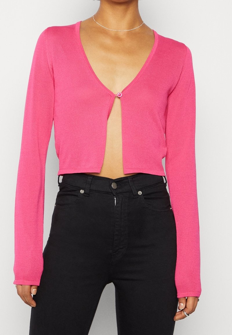 Femme portant un cardigan court à manches longues rose vif, boutonné d'un seul bouton, et un pantalon noir taille haute.