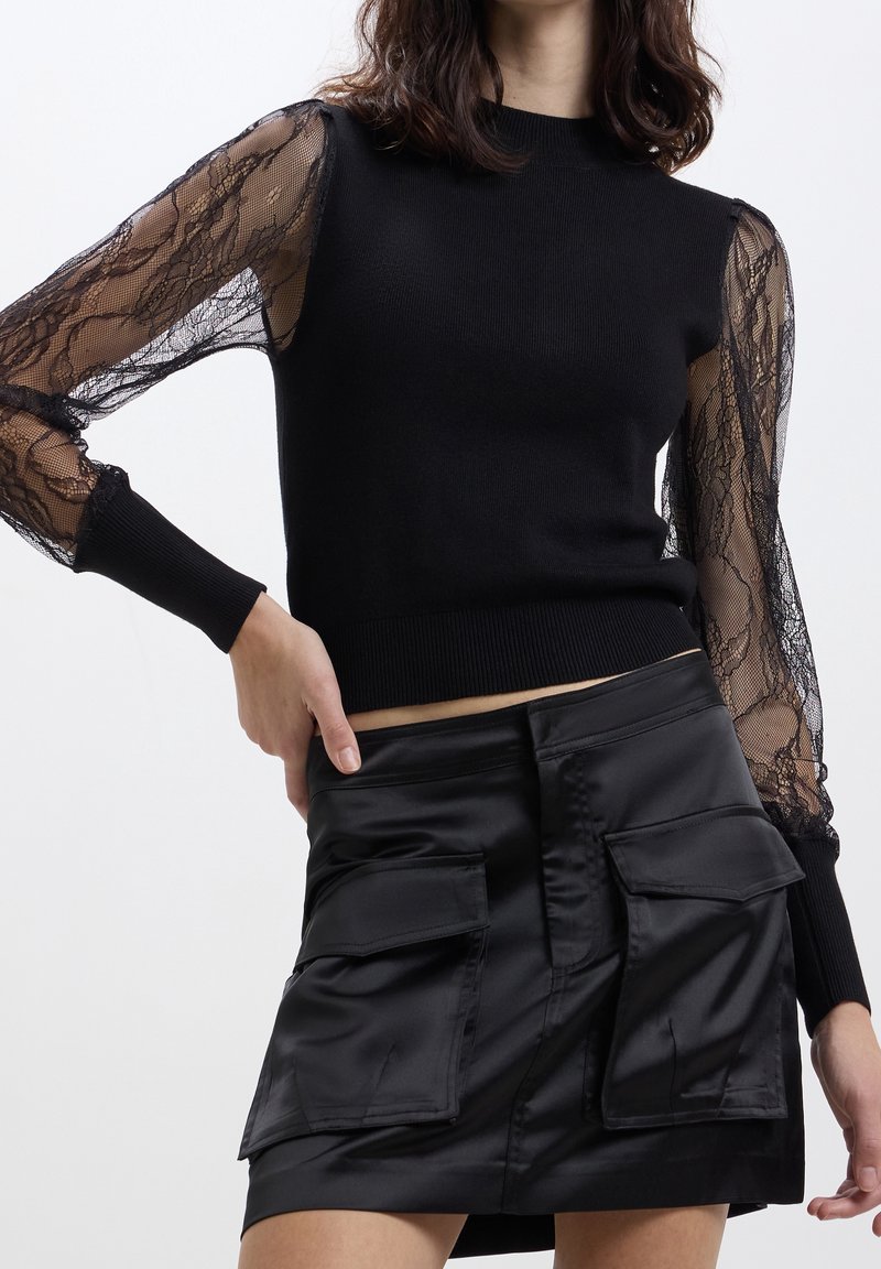 Femme portant un haut noir avec des manches en dentelle transparente et une jupe en satin noire avec de grandes poches avant sur un fond uni.