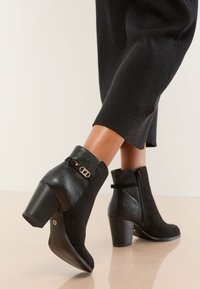 Ghete negre de gleznă, realizate dintr-o combinație de piele întoarsă și piele texturată. Au un toc gros și o curea cu accent auriu. Se remarcă închiderea cu fermoar.