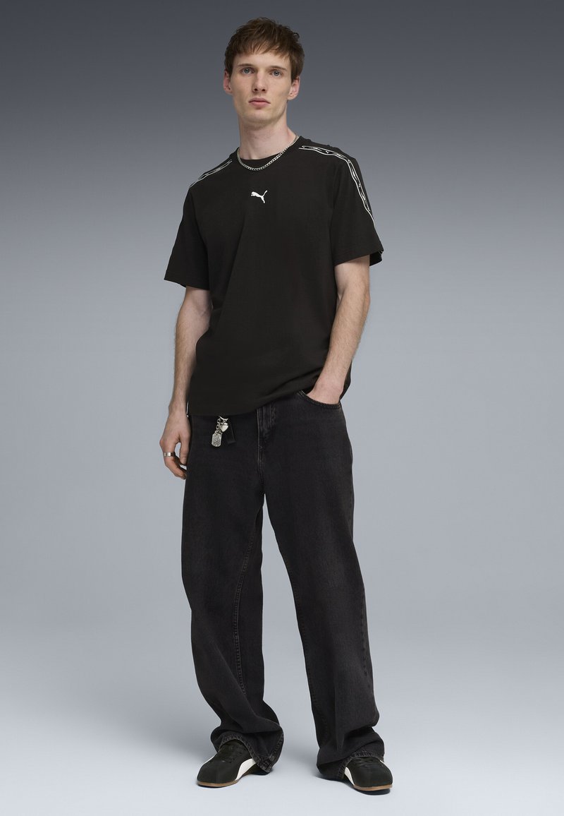 Νεαρός άντρας στέκεται φορώντας μαύρο t-shirt Puma, φαρδιά μαύρα τζιν με μπρελόκ, μαύρα αθλητικά παπούτσια και ασημένιο κολιέ αλυσίδα μπροστά σε γκρι φόντο.