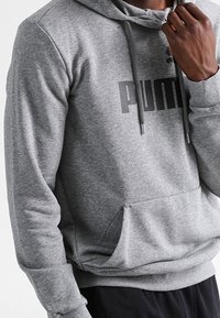 Šedá mikina s kapucí, klokaní kapsou, stahovací šňůrkou a černým logem na hrudi. Vyrobena z měkké, texturované látky.