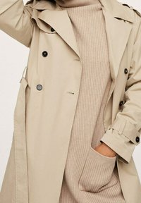 Trench coat beige con design doppiopetto, bottoni neri, colletto a punte e maniche arrotolate, indossato sopra un vestito lungo beige a coste.