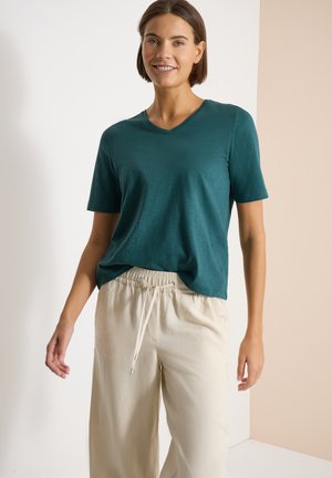 Žena se usmívá, má na sobě tyrkysové tričko s krátkými rukávy a výstřihem do V a béžové kalhoty se stahovací šňůrkou, stojí na neutrálním dvoubarevném pozadí.