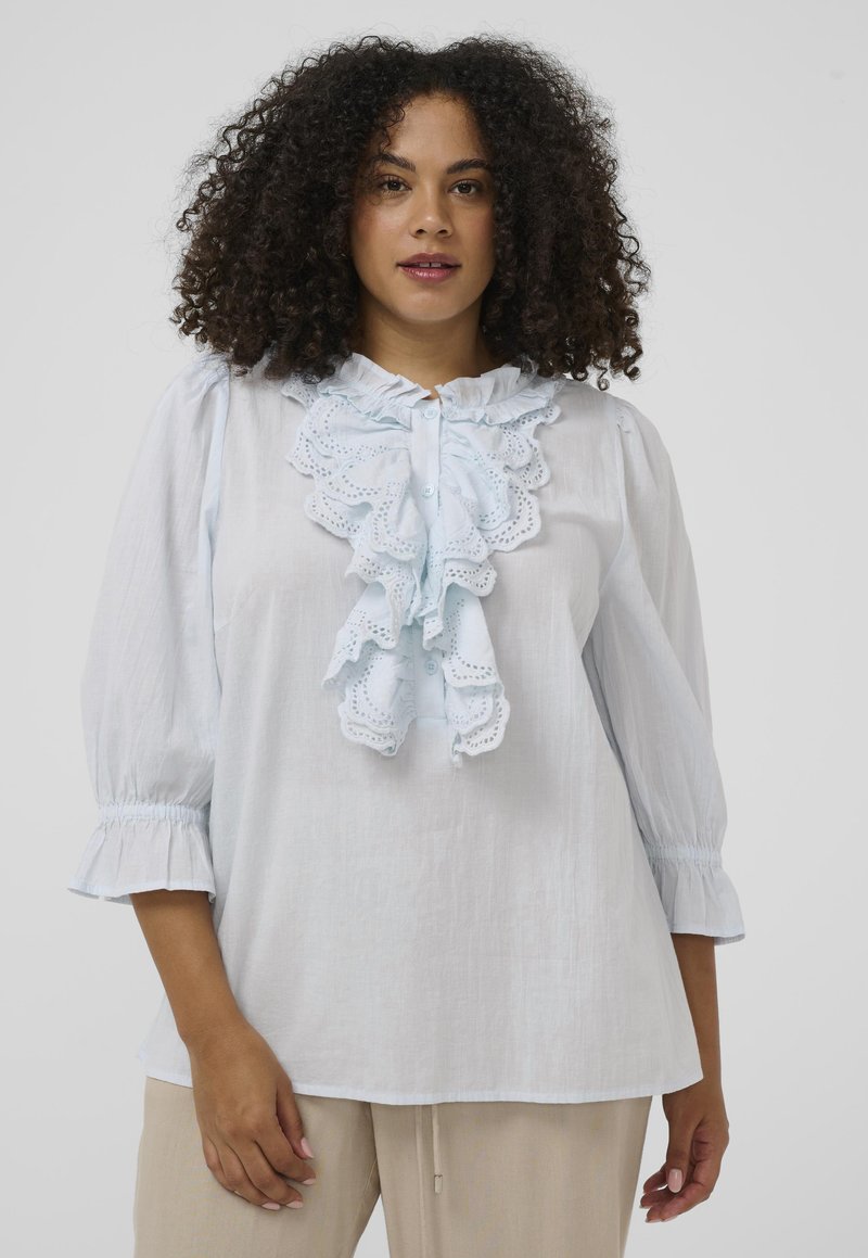 Femme aux cheveux bouclés portant un chemisier bleu clair avec un devant en dentelle à volants et des manches trois-quarts, associée à un pantalon beige, sur un fond uni.