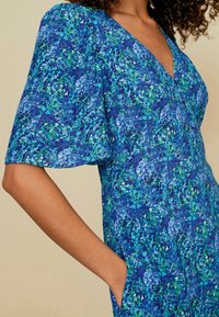 Robe à motif bleu et vert avec un décolleté en V, des manches bouffantes et des poches. Le matériau a une finition texturée avec des éclaboussures abstraites.