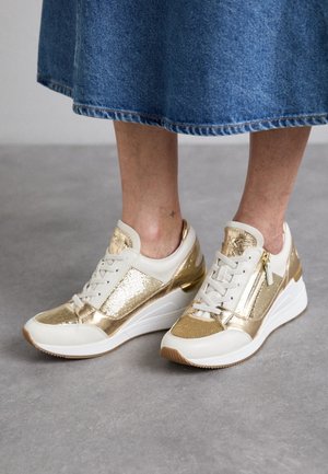 Pieds portant des baskets métalliques blanches et dorées avec une fermeture éclair latérale, associées à une jupe en jean bleue, sur un fond de sol gris.