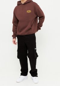 Sudadera marrón con un gráfico amarillo de "MULTIPLY", bolsillo frontal, combinada con pantalones cargo negros con bolsillos laterales y zapatillas blancas.