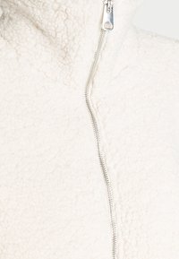 Läheltä otettu kuva valkoisen fleece-takin kankaasta, jossa hopeinen vetoketju on osittain auki.