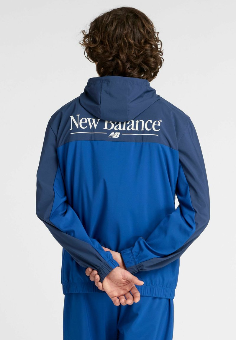 Veste zippée bleue avec capuche ; présente un panneau supérieur navy contrastant et le logo "New Balance". Poignets et ourlet élastiques. Texture lisse.