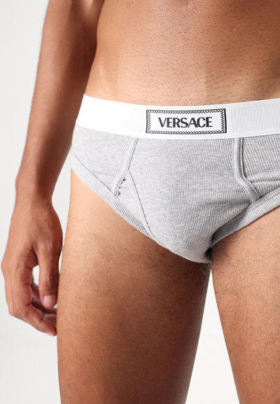 Grijze geribde boxershorts met een witte elastische tailleband met een zwart 'VERSACE' logo etikette. Close-up opname die de pasvorm en stoftextuur benadrukt.