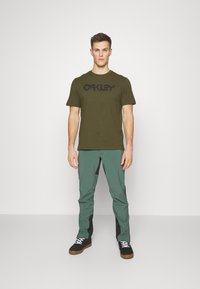 Olivengrøn T-shirt med sort "Oakley" logo, parret med teal bukser med sorte detaljer og sorte sneakers med hvide snørebånd.