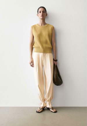 Vrouw staand tegen een witte muur, draagt een mouwloze mosterdgele trui, lichte losse broek met gestrikte manchetten, zwarte sandalen, en houdt een donkere tas vast.