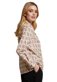 Weiße Bluse mit einem verspielten Muster, das rote und schwarze Illustrationen zeigt. Lange Arme mit Knopfmanschetten und einer lässigen Passform.