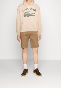 Felpa beige con cappuccio e logo Lacoste verde, abbinata a pantaloncini marroni e sneakers scure con suole crema. Calzini bianchi visibili.