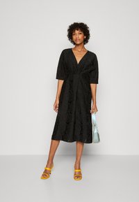 Vestido midi negro con textura de patrón de rosas, escote en V y mangas hasta el codo. Combinado con sandalias de tacón amarillas y un bolso azul claro.