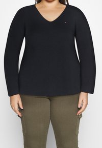 Femme portant un pull noir à col en V et manches longues, ainsi qu'un pantalon cargo olive, debout avec les bras détendus le long du corps.