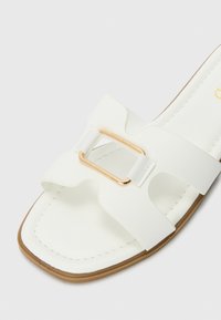White slide sandal featuring a smooth faux leather upper, curved design, wide straps, and a gold-tone rectangular buckle accent.