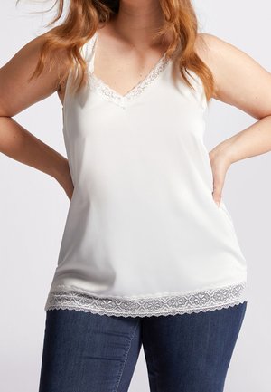 Woman wearing white lace-trimmed sleeveless top and dark blue jeans, standing with hands on hips against plain background.
