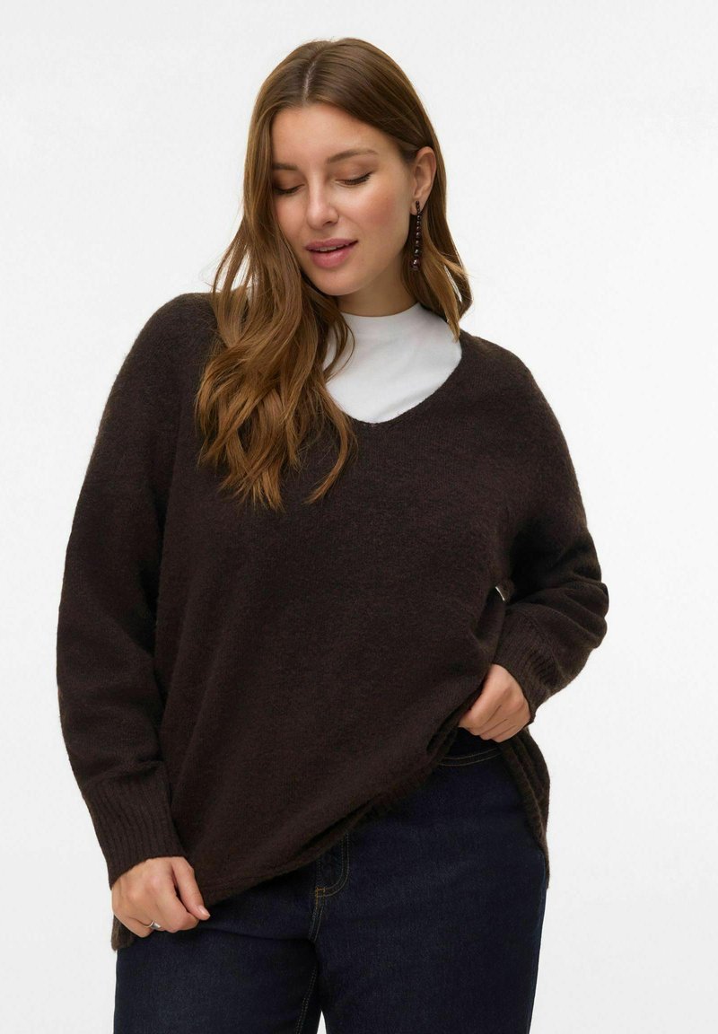 Vrouw met lang bruin haar, gekleed in een donkerbruine V-halstrui over een witte top, terwijl ze de onderkant van de trui vasthoudt, staand tegen een witte achtergrond.