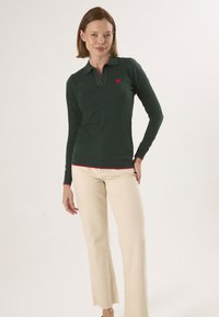Donkergroene lange mouwen polo met een kraag en rode hartborduursel, gecombineerd met lichtbeige vloeiende broek. Glad materiaal, aansluitend ontwerp.