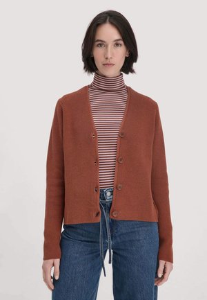 Donna che indossa un cardigan a bottone color ruggine sopra un dolcevita a righe marroni e lavanda, abbinato a jeans blu a vita alta, su uno sfondo neutro.