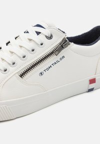 Witte leren sneaker met een gestructureerd oppervlak, voorzien van een ritssluiting aan de zijkant, witte veters en een rode, blauwe en witte accent op de hiel.