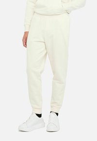 Pantalones de chándal color crema con cintura ajustable, piernas ajustadas y bolsillos laterales, combinados con zapatillas blancas. Textura suave y diseño minimalista.