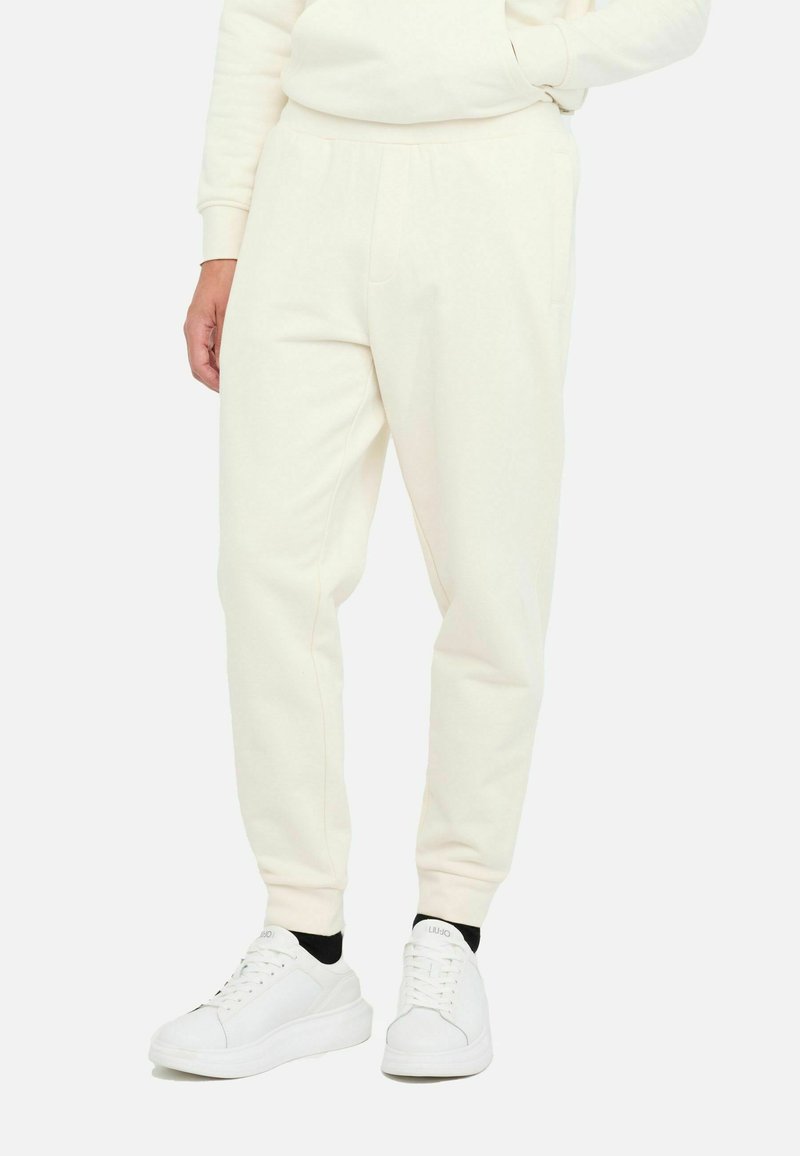 Cremefarbene Sweatpants mit verstellbarem Bund, tapered Beinen und seitlichen Taschen, kombiniert mit weißen Sneakers. Weicher Stoff und minimalistisches Design.